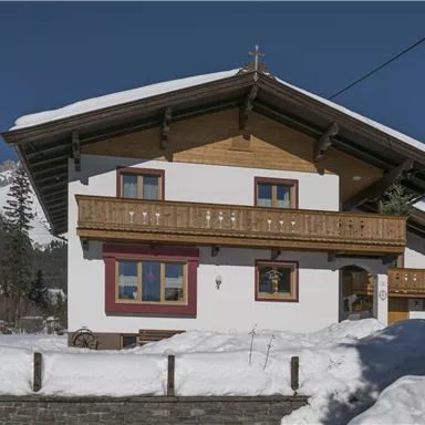 A charming house in the snow with a wooden balcony. In the background, snow-covered mountains and a blue sky can be seen.