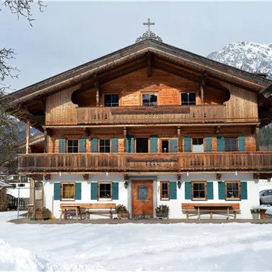 A traditional wooden house in Bavarian style, surrounded by snow. Snow-covered mountains can be seen in the background.
