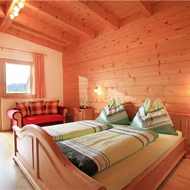 A bright bedroom with wooden paneling and a generous window. The room is cozily furnished with a double bed and a seating area.