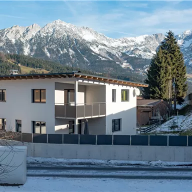 Ein modernes Haus in den Bergen, umgeben von schneebedeckten Landschaften. Im Hintergrund liegen beeindruckende Berge unter einem klaren Himmel.