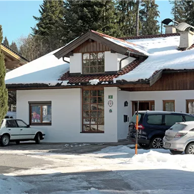 A white house with a red roof, surrounded by snow and trees. Parked cars are in front of the building.