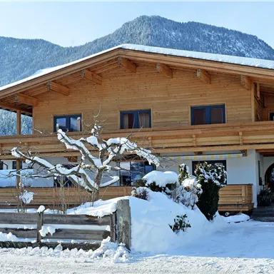 Ein schönes Holzhaus im Schnee mit einem großen Balkon. Im Hintergrund erheben sich schneebedeckte Berge unter einem blauen Himmel.