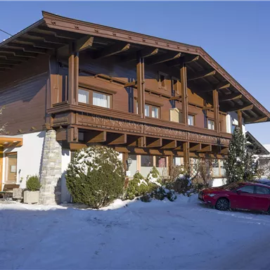 Ein traditionelles Holzhaus in einem verschneiten Bereich. Im Vordergrund steht ein rotes Auto vor dem Gebäude.