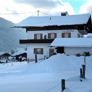 Ein schneebedecktes Haus in einer ruhigen winterlichen Landschaft. Im Hintergrund sind schneebedeckte Berge und weitere Gebäude zu sehen.