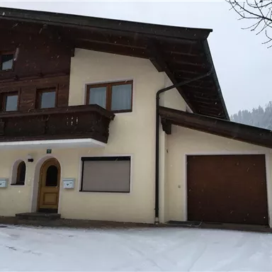 A traditional alpine house with a wooden balcony and a garage door. Snow covers the ground and surrounds the building.