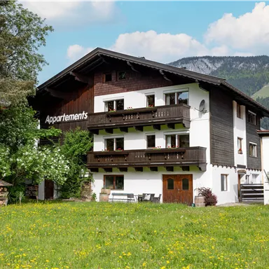 Ein gemütliches Haus mit Balkonen und einem schönen Garten. Die Umgebung zeigt grüne Wiesen und Berge im Hintergrund.