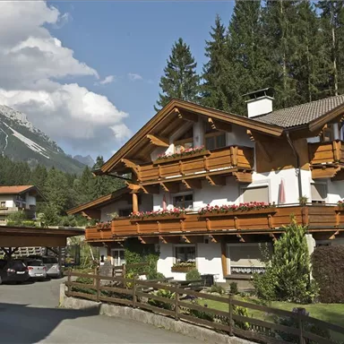 Ein traditionelles Chalet umgeben von Bäumen und Bergen. Die Balkone sind mit Blumen geschmückt, und die Umgebung ist ruhig und malerisch.