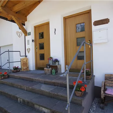 Ein einladender Hauseingang mit Holztreppe und zwei Türen. Bunte Blumen und eine Bank im Eingangsbereich verleihen einen freundlichen Eindruck.