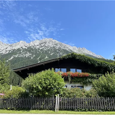 Ein charmantes Haus umgeben von üppigem Grün und bunten Blumen. Im Hintergrund ragen majestätische Berge under einem klaren blauen Himmel empor.