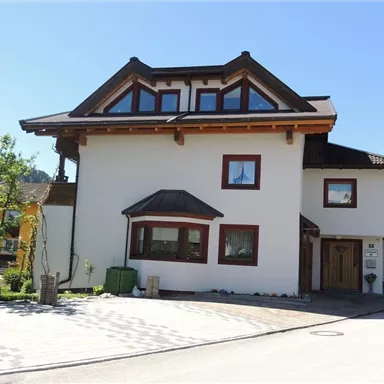 A modern house with a one-story design and a gable roof. The front yard is well-kept and surrounded by trees.