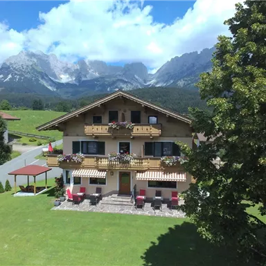 A cozy house in the mountains with blooming balconies. In the background, majestic mountains and a clear blue sky can be seen.