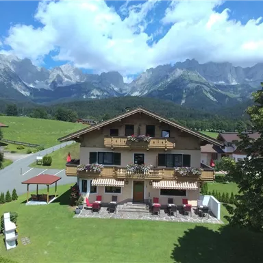 A beautiful chalet with balconies and flowers. Impressive mountains and a blue sky can be seen in the background.