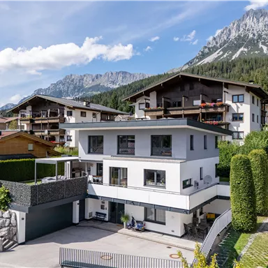 A modern house with two floors in a picturesque mountain landscape. In the background, high mountains and other residential buildings are visible.