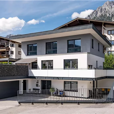 A modern house with large windows and a clear parking lot. In the background, mountains and a beautiful landscape are visible.