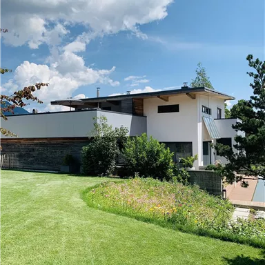 Ein modernes Haus mit einer grünen Rasenfläche und bunten Blumen im Vordergrund. Der Himmel ist blau mit einigen Wolken.