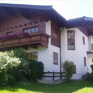 A beautiful house in alpine style with a wooden balcony and blooming flowers. Green bushes surround the building and the sky is clear.