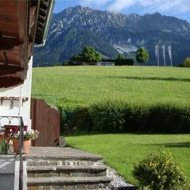 A picturesque view of a green meadow with mountains in the background. In the foreground, there is a terrace with chairs and a small garden.
