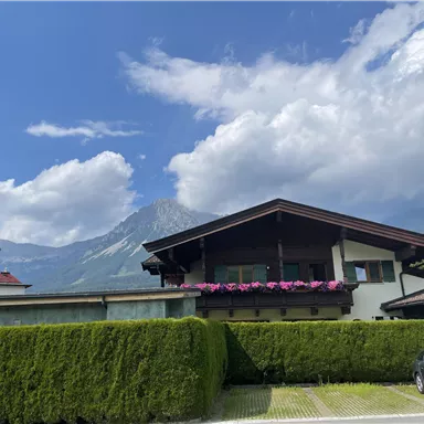 A beautiful house with a garden and colorful flowers on the balcony. In the background, mountains and a blue sky can be seen.