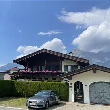 A beautiful house with balconies and colorful flowers. In the background, mountains and a clear sky can be seen.