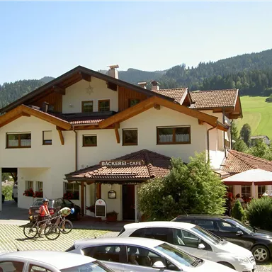 A traditional building with a café and a bakery, surrounded by green hills. In front of the house, there are some parking spaces and bicycles.