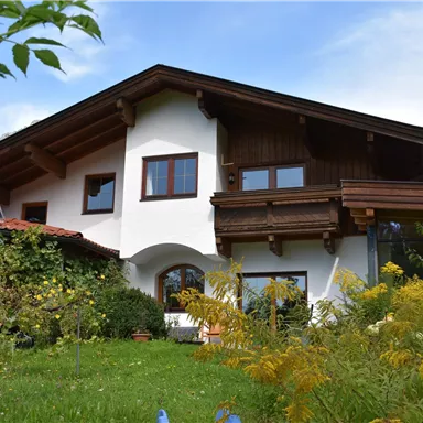 A charming house with wood siding and large windows in a green garden. Gentle hills are visible in the background.