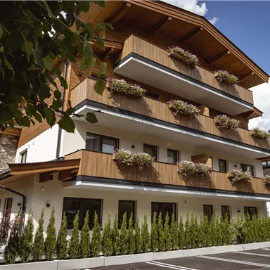A modern building with wooden-clad balconies and many flowers. It is situated in a quiet environment with mountains in the background.