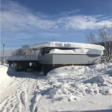 Ein modernes Haus inmitten von tiefem Schnee. Der Himmel ist klar und blau, während eine dicke Schneeschicht auf dem Dach liegt.
