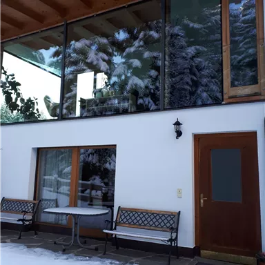 A modern house with large windows and a wooden ceiling. Outside, there is snow, and there is a small terrace with a table and benches.