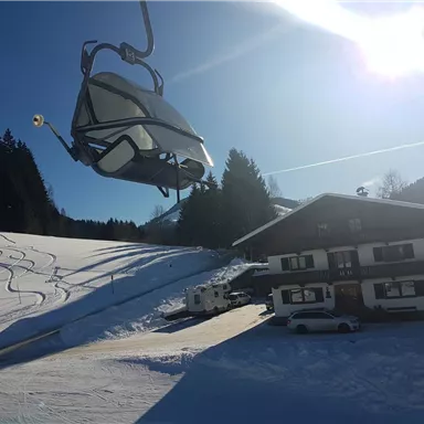 Eine sonnige Winterlandschaft mit einem Skilift und verschneiten Hügeln. Im Vordergrund steht ein gemütliches Berghaus.