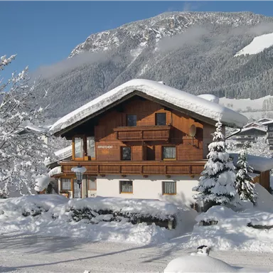 A cozy wooden house in the snow, surrounded by snow-covered trees. In the background, majestic mountains rise under a clear blue sky.