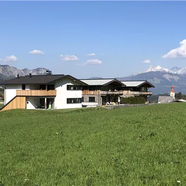 A modern building is situated in a green landscape. In the background, mountains and a small church tower are visible.