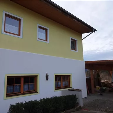 A modern two-story house with a yellow and white facade. In the foreground, there are windows with wooden frames and an adjacent covered area visible.