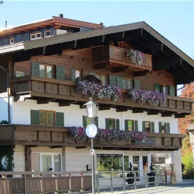 Ein traditionelles Chalet mit Holzbalkonen und bunten Blumen. Es befindet sich in einer malerischen Umgebung mit grünen Bäumen.