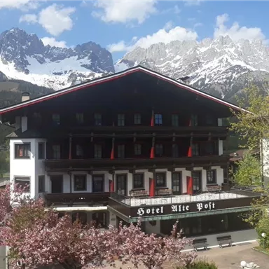 A traditional Alpine hotel with a blooming garden in the foreground. In the background, majestic mountains and a bright blue sky can be seen.