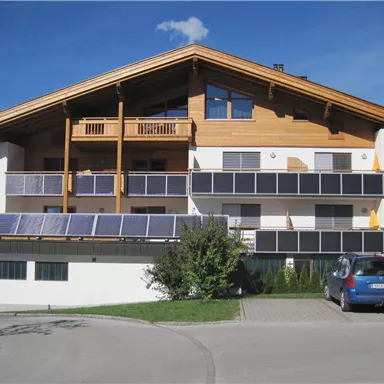 Ein modernes Gebäude mit Holzdetails und mehreren Balkonen. Es gibt Solarpanels auf dem Dach und einen Parkplatz vor dem Haus.