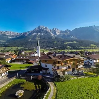 A picturesque village view with traditional houses and a church, surrounded by green meadows and mountains. The sky is clear and blue, which emphasizes the idyllic landscape.