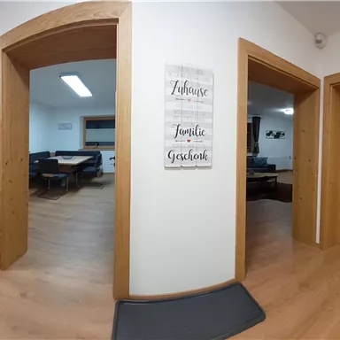 Ein helles Zimmer mit mehreren Türen und einem Holzfußboden. An der Wand hängt ein dekoratives Schild mit positiven Sprüchen.