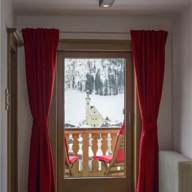 A view from a room with red curtains onto a snowy outdoor area. In the background, a church and a picturesque landscape can be seen.