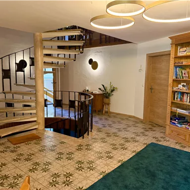 An inviting entrance area with a curved staircase and a bookshelf. The floor is designed with a pretty pattern, and there is plenty of daylight.