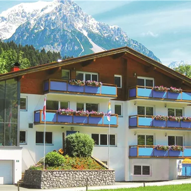 A modern house with several balconies, surrounded by green trees. In the background, majestic, snow-capped mountains rise.