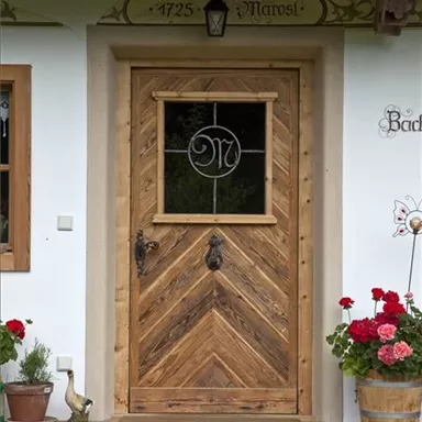 A rustic wooden door with an elegant window in the middle. In front of the door, red geraniums are blooming and a small decorative object is placed in the garden.