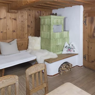 A rustic room with wood paneling and a cozy seating area. In the corner stands a traditional stove in green color.