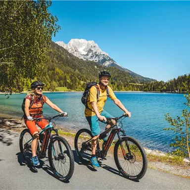 Zwei Radfahrer fahren an einem klaren blauen See entlang, umgeben von grünen Bäumen und Bergen. Der Himmel ist sonnig und blau.