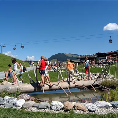 Children are playing on a wooden bridge over a small waterway. In the background, mountains and a cable car can be seen.