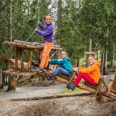 Three children are playing on a wooden cart in the forest. They are joyful and wearing colorful clothes.
