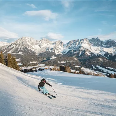A skier goes down the snowy slope, surrounded by majestic mountains. The sky is clear and the landscape is wintery beautiful.