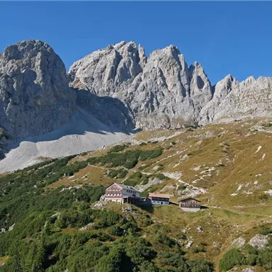 Eine beeindruckende Berglandschaft mit steilen Felsen und grünem Gelände. In der Mitte steht ein Berggasthaus, umgeben von Natur.