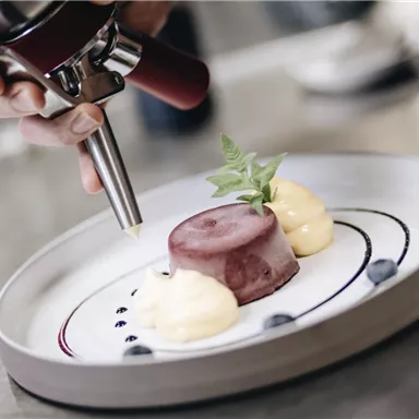 A person is decorating a dessert on a white plate. The dessert consists of a red jelly and two dollops of cream, decorated with a mint leaf.