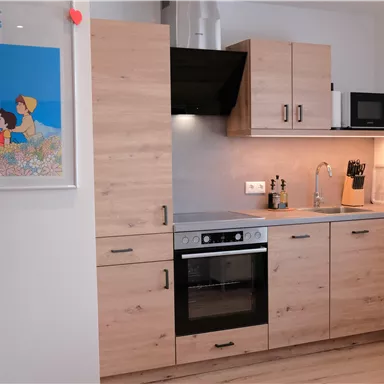 A modern kitchen with wooden cabinets, a stove, and a microwave. A colorful picture of "Heidi" hangs on the wall.