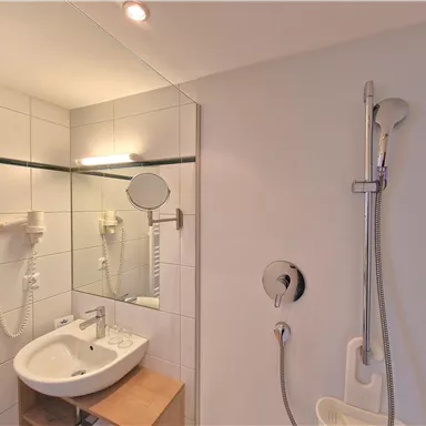 A modern bathroom with a shower and a sink. The walls are tiled with white tiles and there is a mirror.